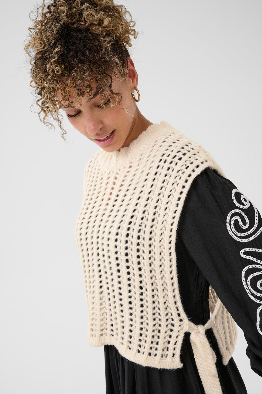 Woman wearing a beige crochet vest over a black long-sleeve shirt with white patterns.