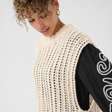 Woman wearing a beige crochet vest over a black long-sleeve shirt with white patterns.