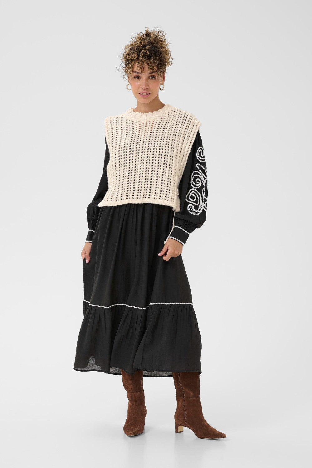 Woman wearing a black dress with a cream knit sweater vest over it on a white background