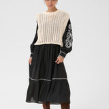 Woman wearing a black dress with a cream knit sweater vest over it on a white background