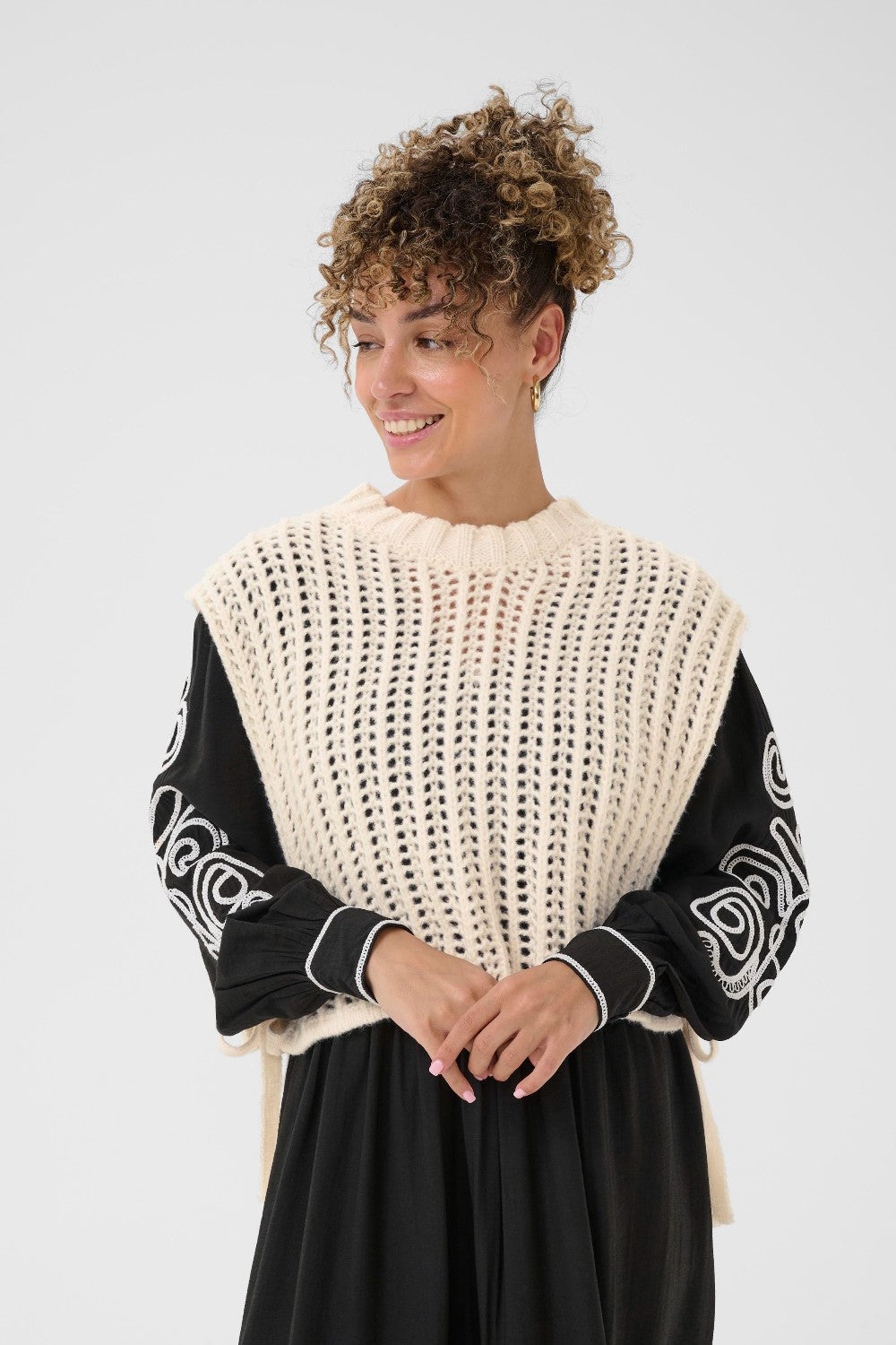 Woman wearing a black and white patterned top with a beige knit vest over it on a plain background