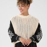 Woman wearing a black and white patterned top with a beige knit vest over it on a plain background
