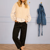 Woman wearing a sleeveless cream sweater over a blouse with black pants standing in a room with a wooden floor and light blue wall.