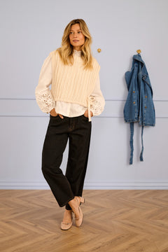 Woman wearing a sleeveless cream sweater over a blouse with black pants standing in a room with a wooden floor and light blue wall.