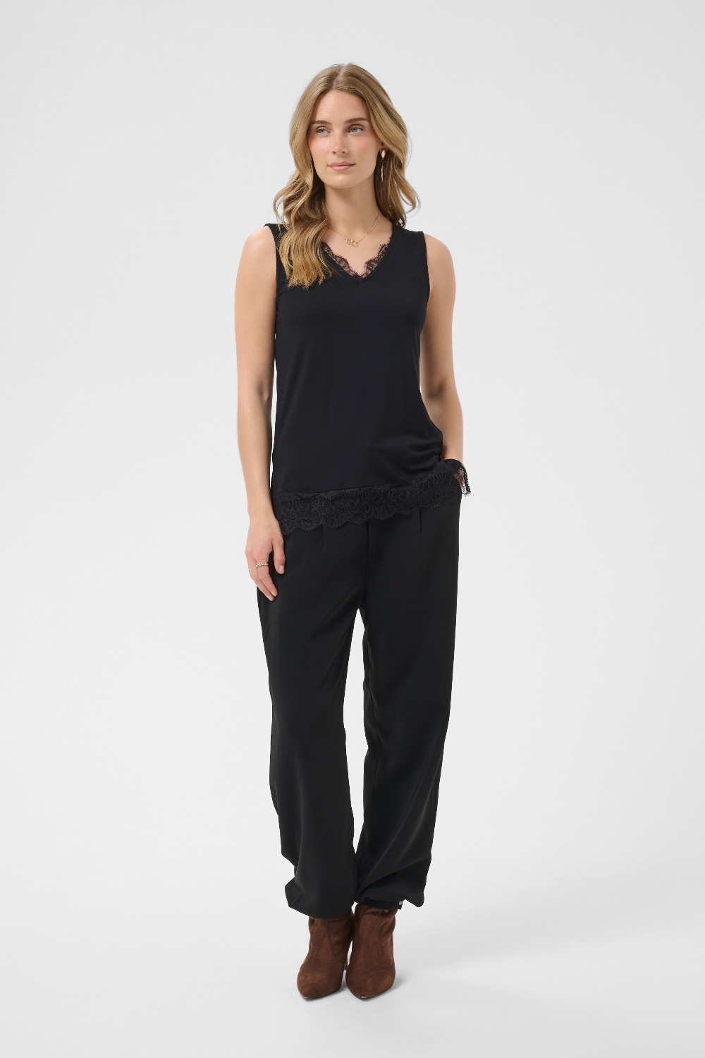 A woman modeling a black sleeveless top with a lace hem and black wide-leg pants, paired with brown ankle boots, against a light gray studio background.