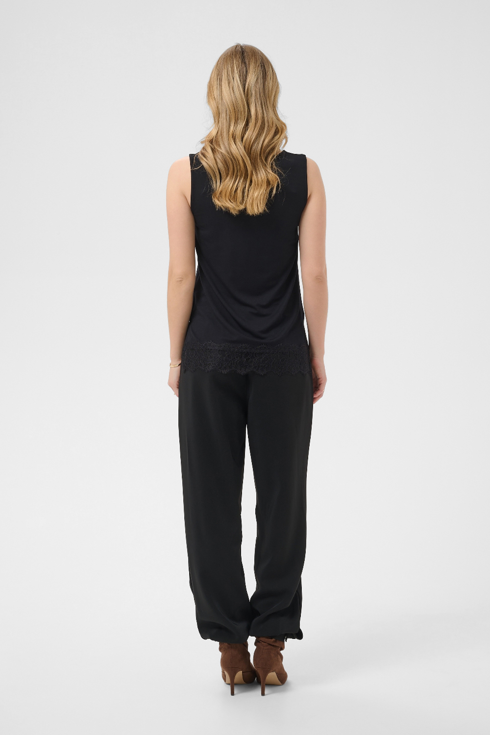 A woman viewed from behind wearing a black sleeveless top and black wide-leg pants, standing against a light gray studio background.