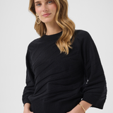 A woman wearing a black 3/4-sleeve knit sweater with a textured fabric, paired with dark blue jeans, against a light gray studio background.