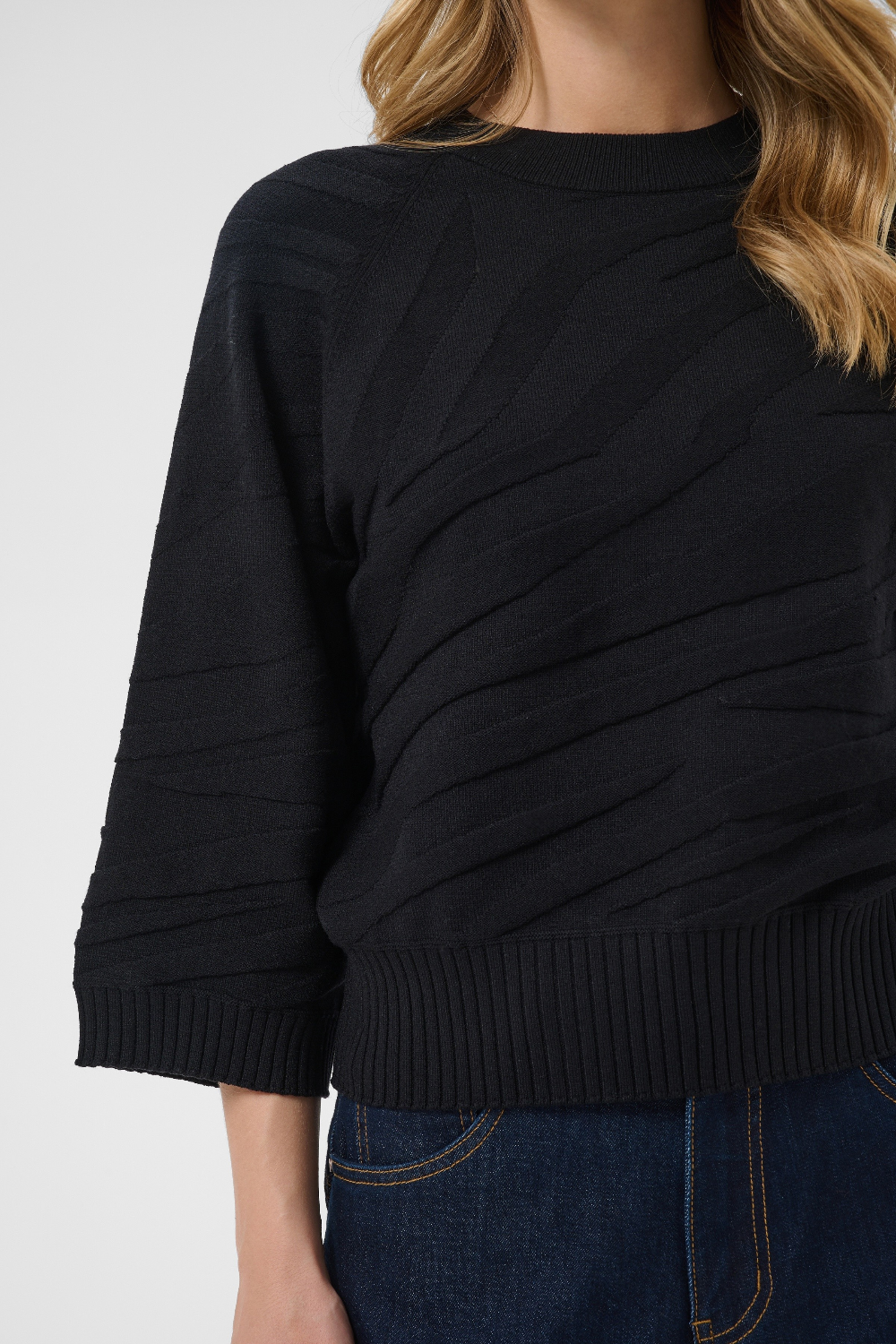 A close-up of a woman wearing a black ribbed knit sweater with diagonal textured paneling on the front, paired with dark blue jeans against a light gray background.