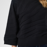 A close-up of a woman wearing a black ribbed knit sweater with diagonal textured paneling on the front, paired with dark blue jeans against a light gray background.