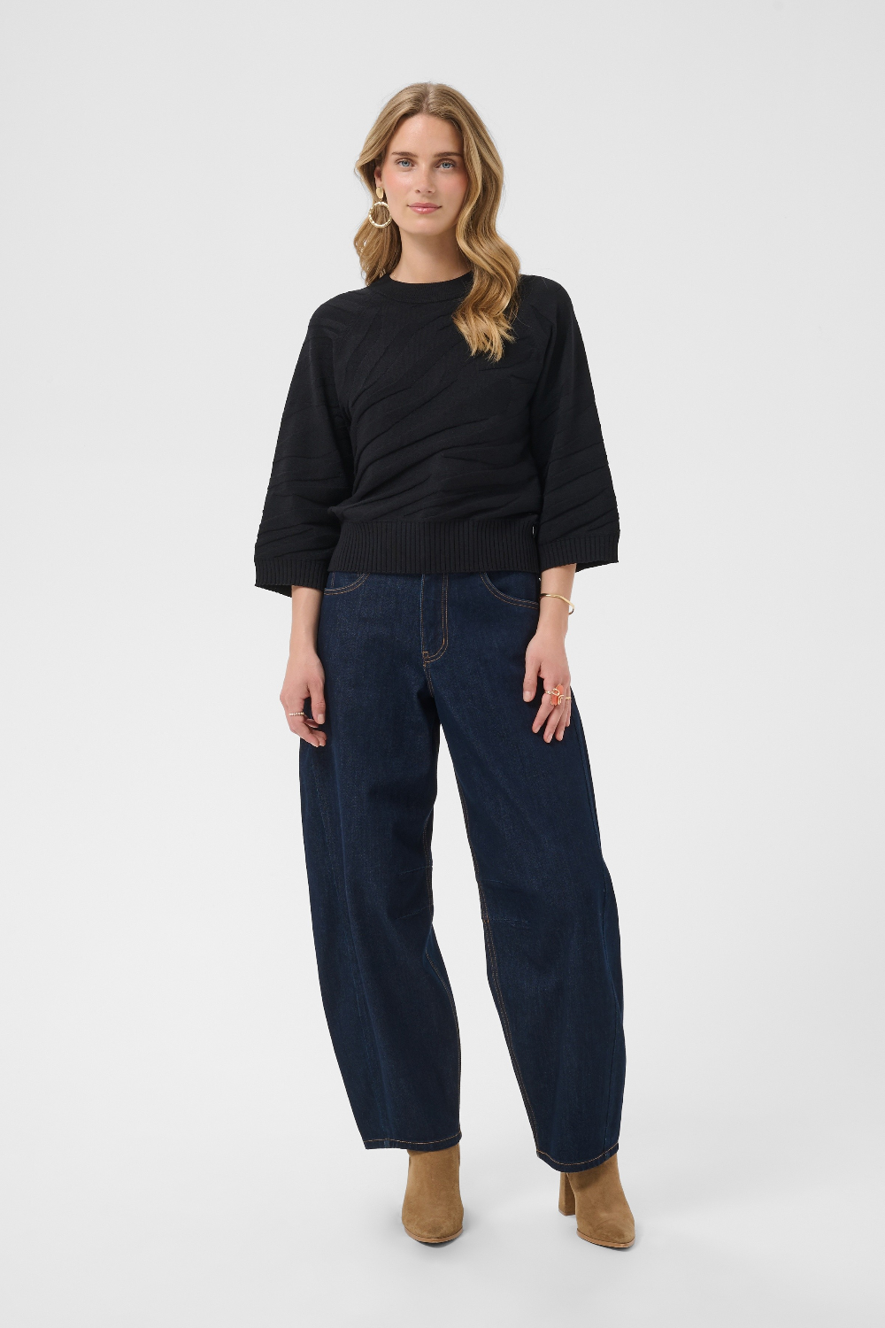 A woman modeling a black 3/4-sleeve top and dark blue wide-leg jeans with tan ankle boots, standing against a light gray studio background.
