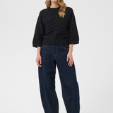 A woman modeling a black 3/4-sleeve top and dark blue wide-leg jeans with tan ankle boots, standing against a light gray studio background.