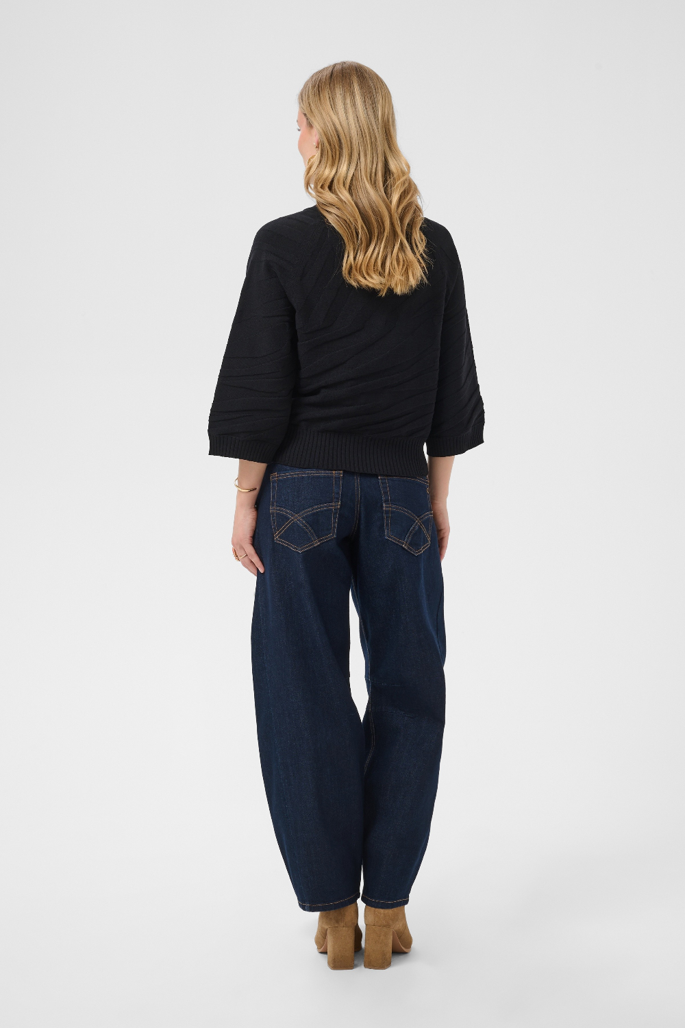 A woman shown from the back wearing a black ribbed knit sweater, dark blue wide-leg jeans with contrast stitching, and tan block-heel boots, against a light gray studio background.