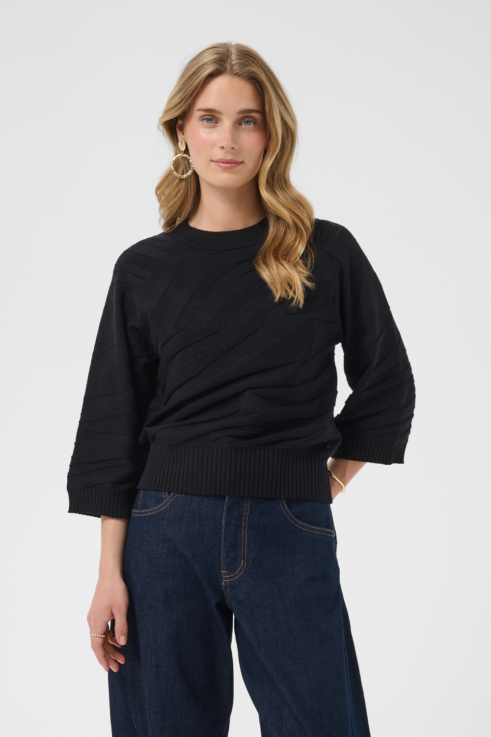 A woman modeling a black 3/4-sleeve knit sweater with a ribbed hem, paired with dark blue jeans, against a light gray studio background.