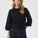 A woman modeling a black 3/4-sleeve knit sweater with a ribbed hem, paired with dark blue jeans, against a light gray studio background.
