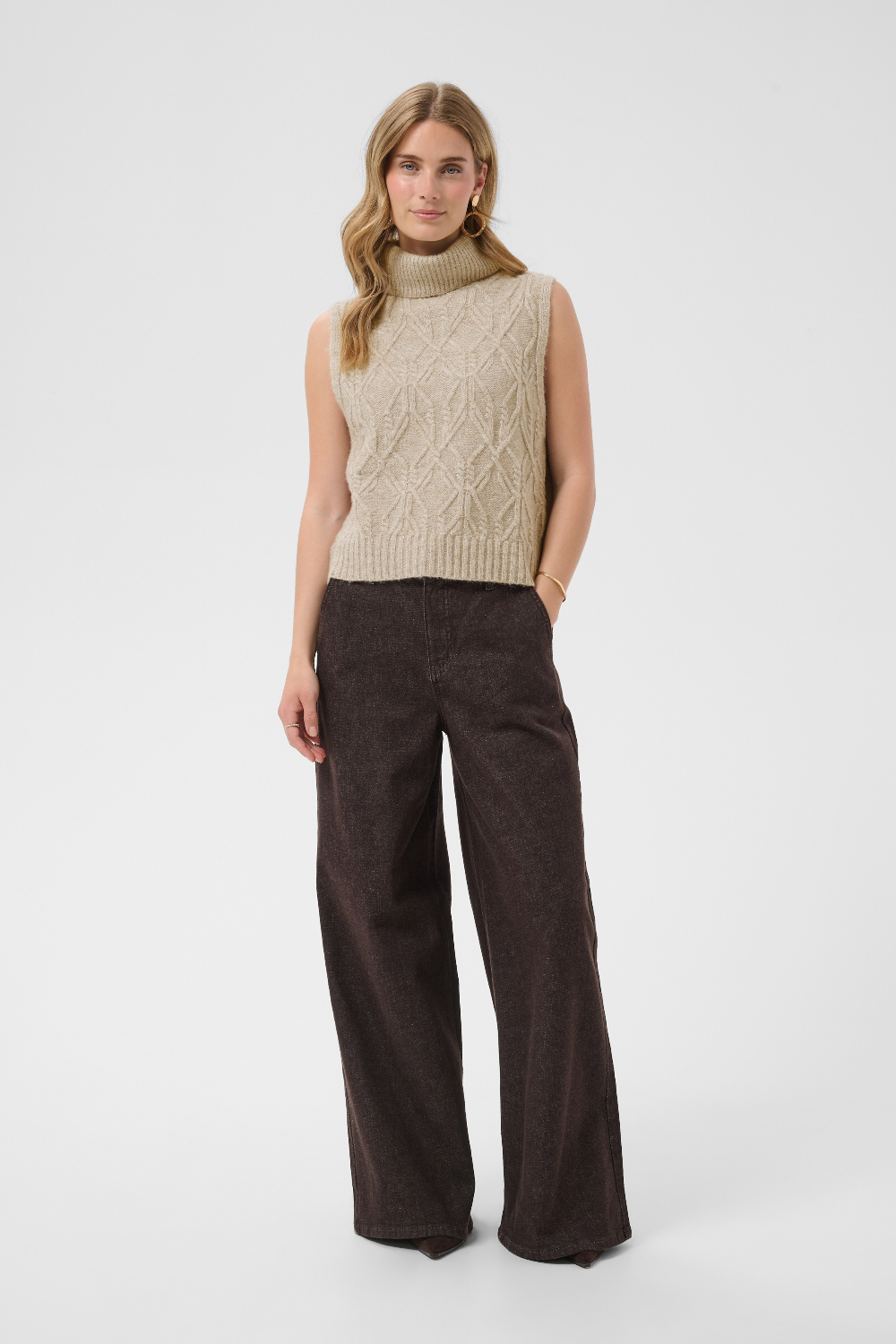 A woman wearing a sleeveless beige knit vest and dark brown wide-leg trousers against a light gray studio background.