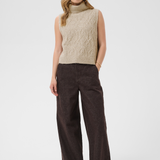 A woman wearing a sleeveless beige knit vest and dark brown wide-leg trousers against a light gray studio background.