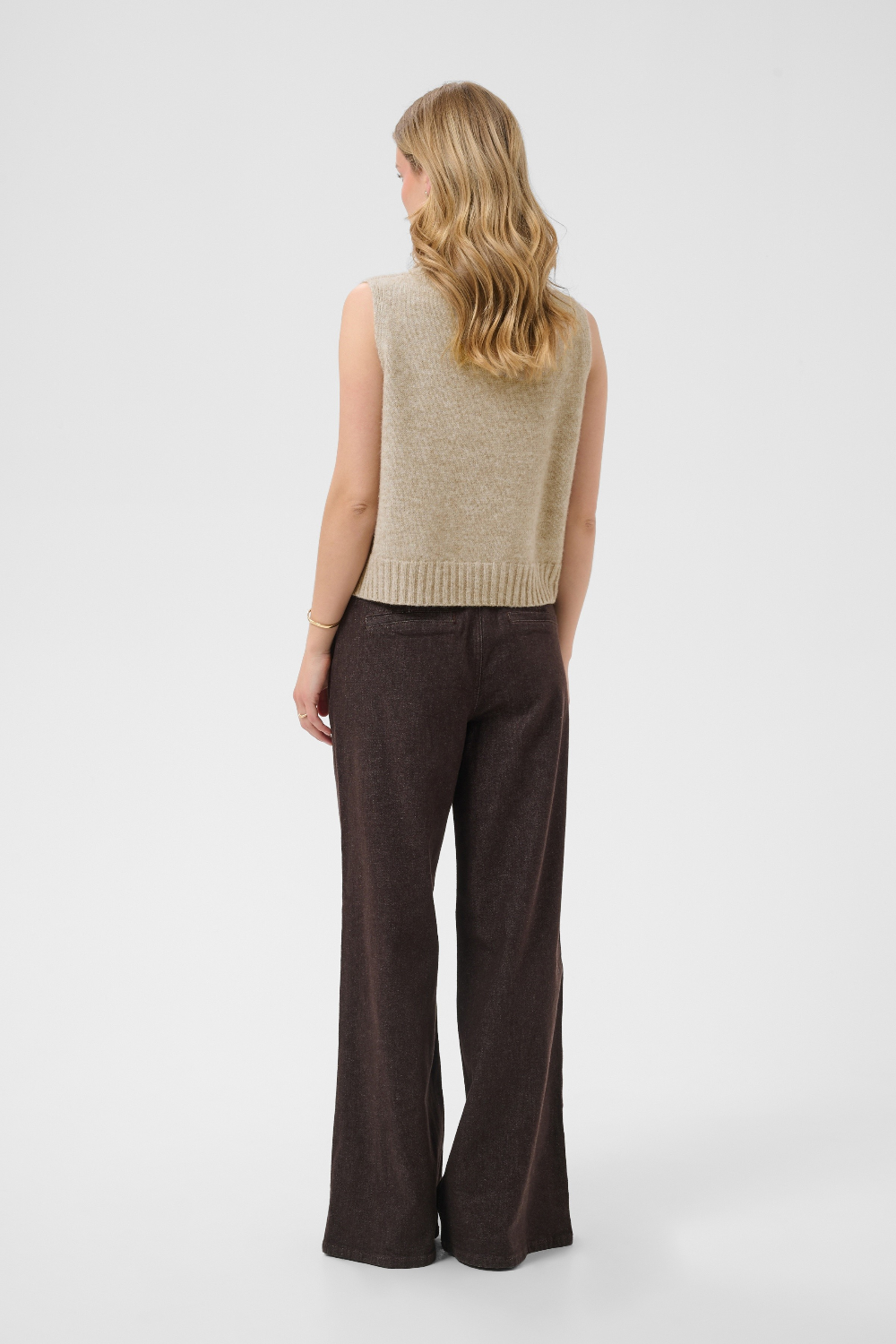 A woman shown from the back wearing a sleeveless beige knit vest and dark brown wide-leg trousers against a light gray studio background.