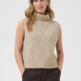 A woman wearing a sleeveless cable-knit mock neck sweater vest in a light beige color, paired with dark brown pants, against a light gray studio background.