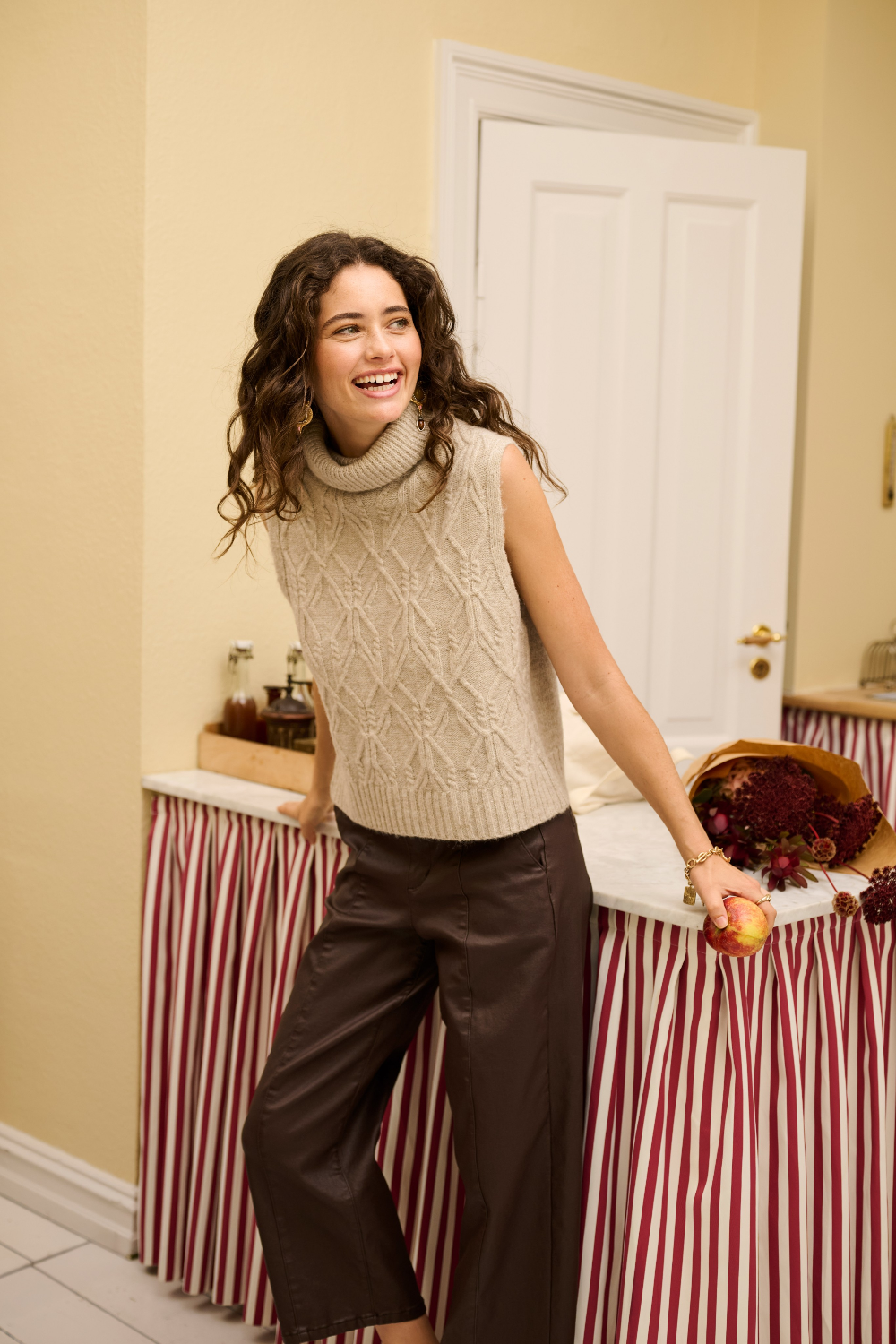 A woman wearing a sleeveless beige knit vest and dark brown wide-leg cropped trousers in a warm indoor setting.