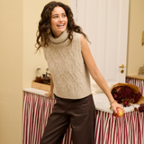 A woman wearing a sleeveless beige knit vest and dark brown wide-leg cropped trousers in a warm indoor setting.