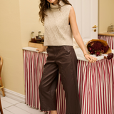 A woman wearing a sleeveless beige knit vest and dark brown wide-leg cropped trousers in a warm indoor setting.