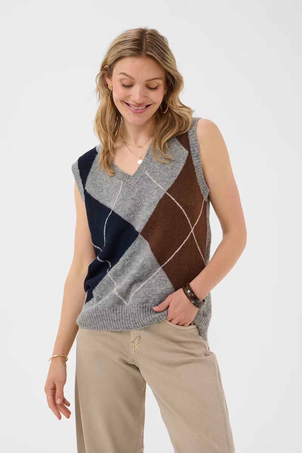 Woman wearing a sleeveless grey sweater featuring a dark blue and brown argyle design with tan pants against a light background.