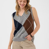 Woman wearing a sleeveless grey sweater featuring a dark blue and brown argyle design with tan pants against a light background.