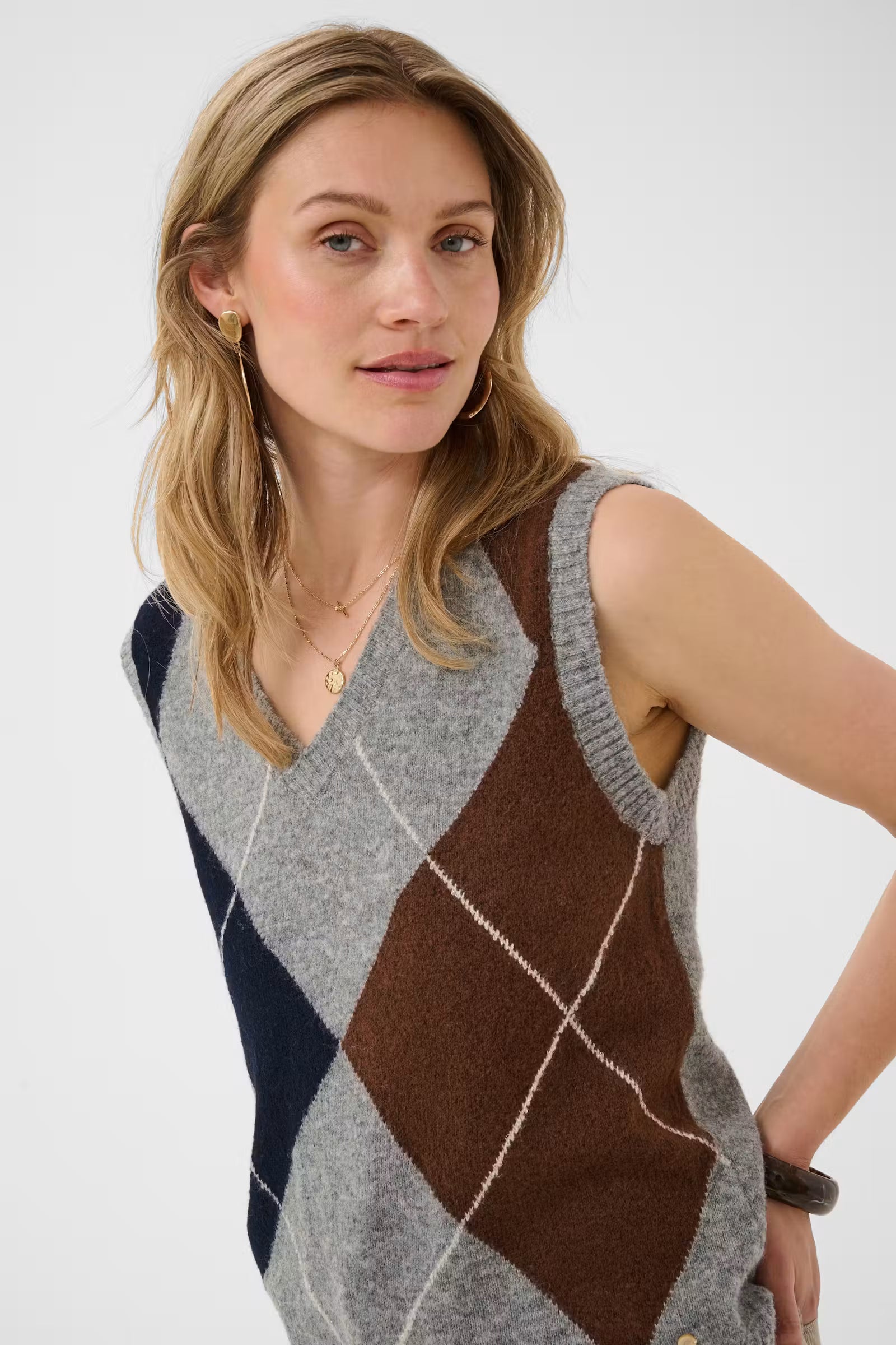 Woman wearing a sleeveless grey sweater featuring a dark blue and brown argyle design against a light background.