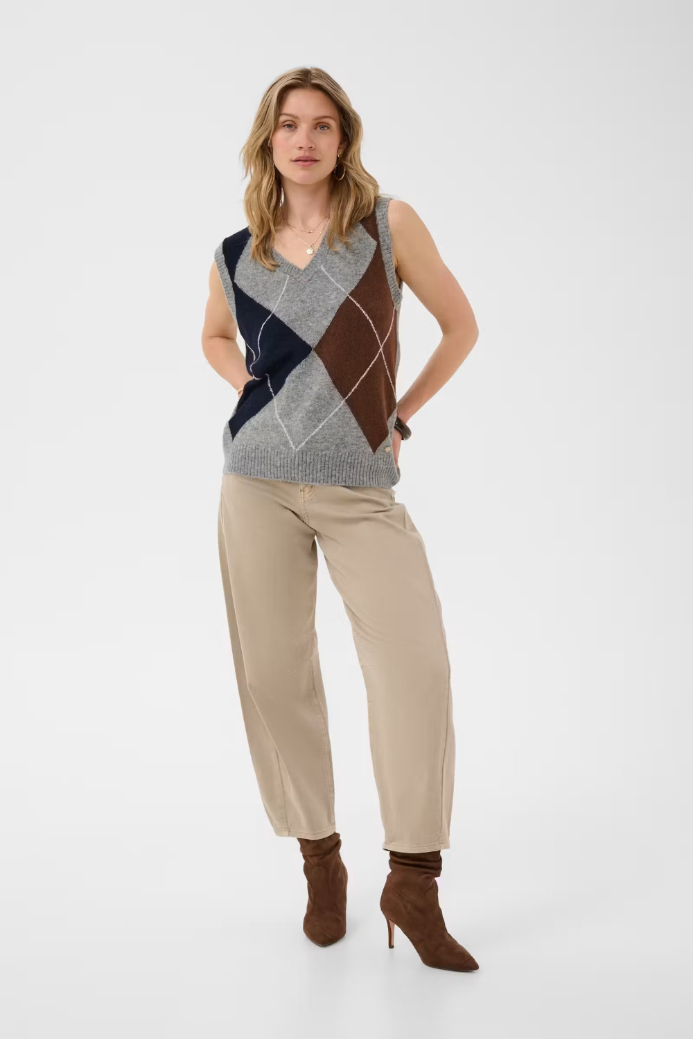 Woman wearing a sleeveless grey sweater featuring a dark blue and brown argyle design with tan pants and brown boots against a light background.