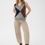 Woman wearing a sleeveless grey sweater featuring a dark blue and brown argyle design with tan pants and brown boots against a light background.