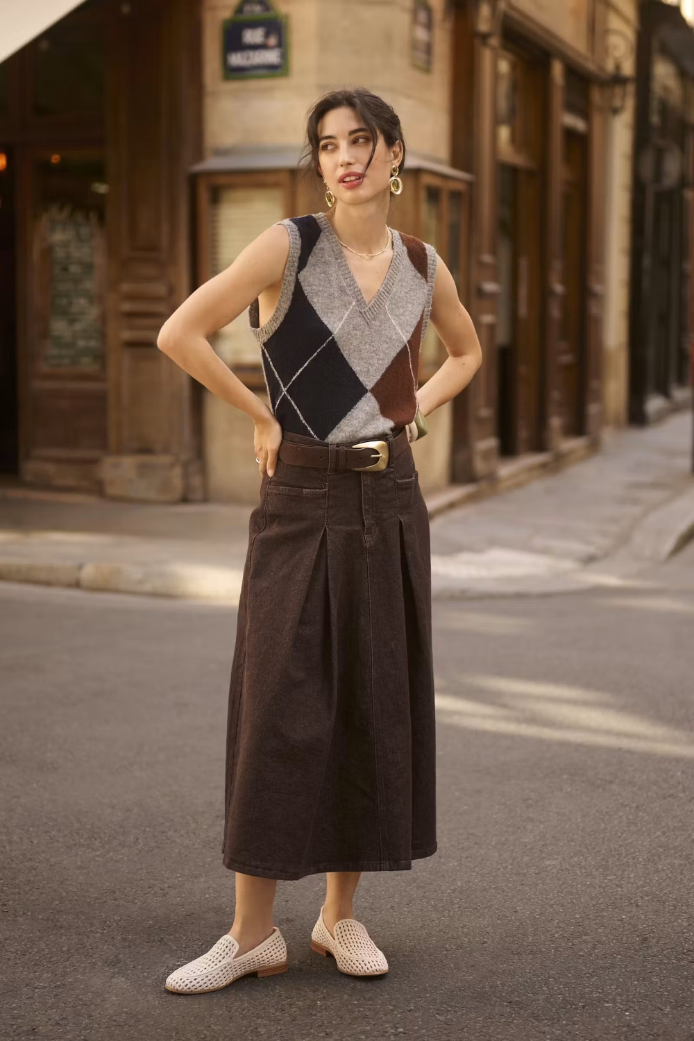 Woman wearing a sleeveless grey sweater featuring a dark blue and brown argyle design with a brown skirt in an outdoor city setting.