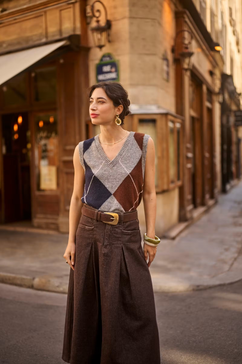 Woman wearing a sleeveless grey sweater featuring a dark blue and brown argyle design with a brown skirt in an outdoor city setting.