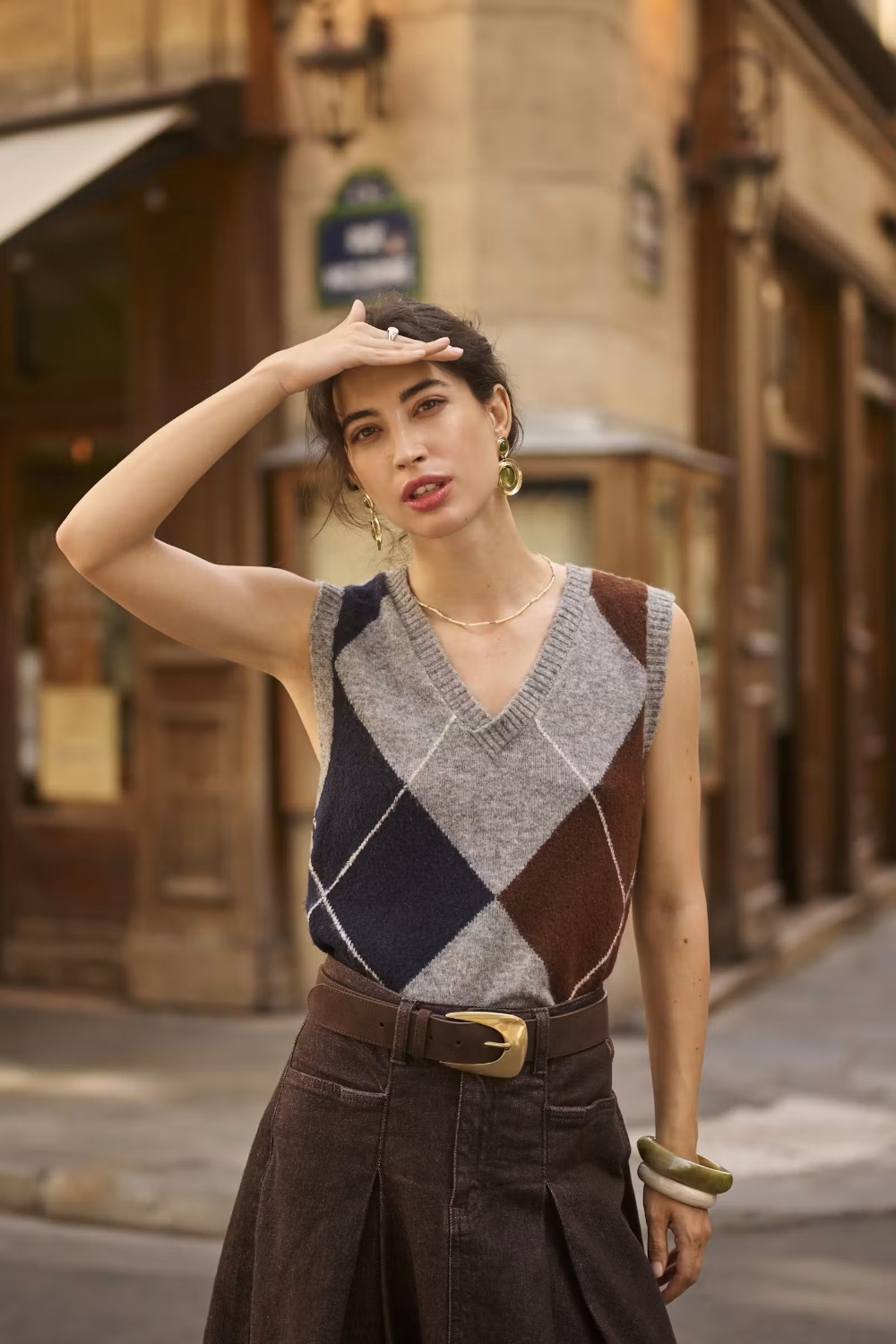 Woman wearing a sleeveless grey sweater featuring a dark blue and brown argyle design with a brown skirt in an outdoor city setting.