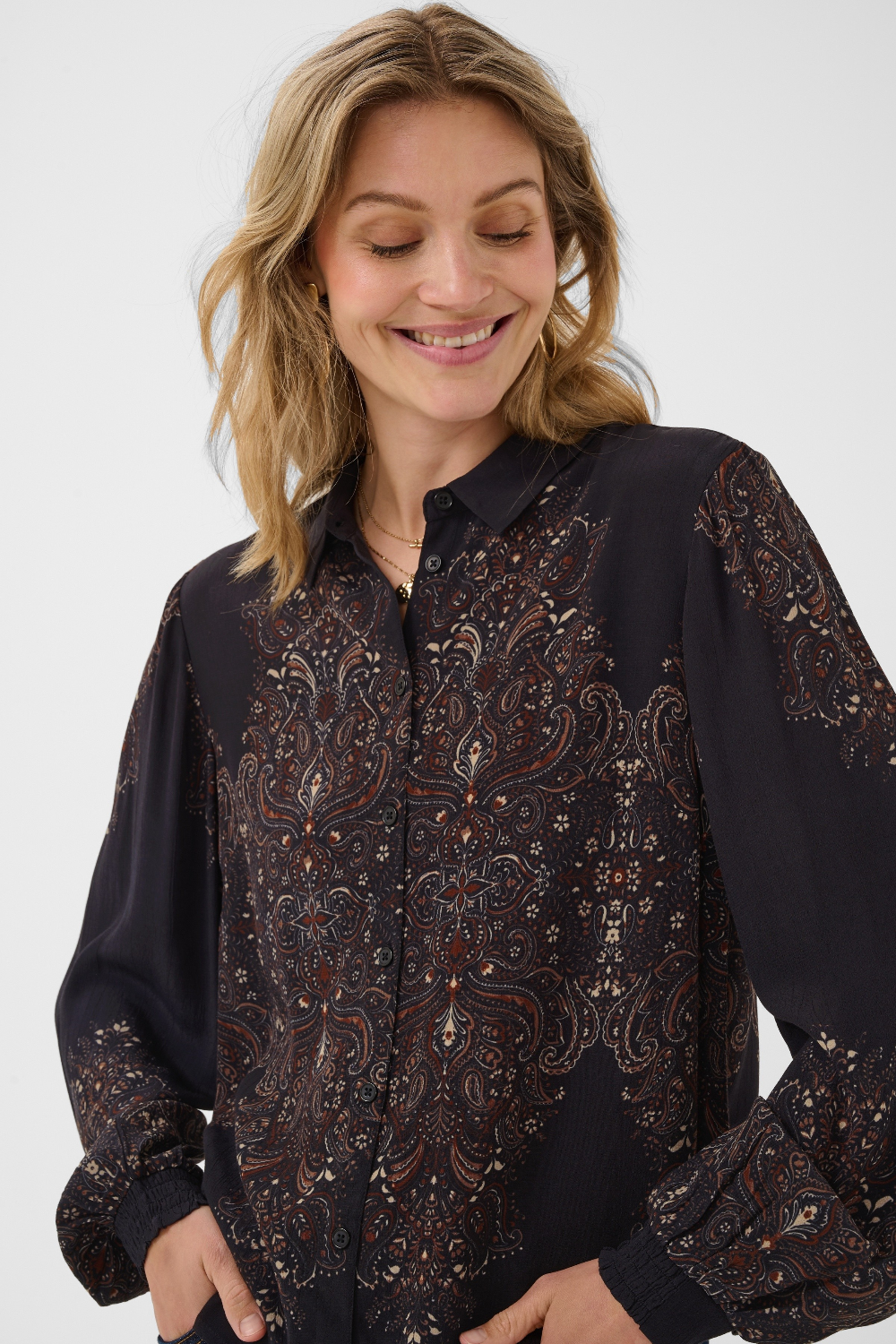 A woman wearing a dark long-sleeve button-up blouse with an intricate brown, beige, and orange paisley pattern, photographed against a light gray studio background.