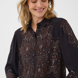 A woman wearing a dark long-sleeve button-up blouse with an intricate brown, beige, and orange paisley pattern, photographed against a light gray studio background.