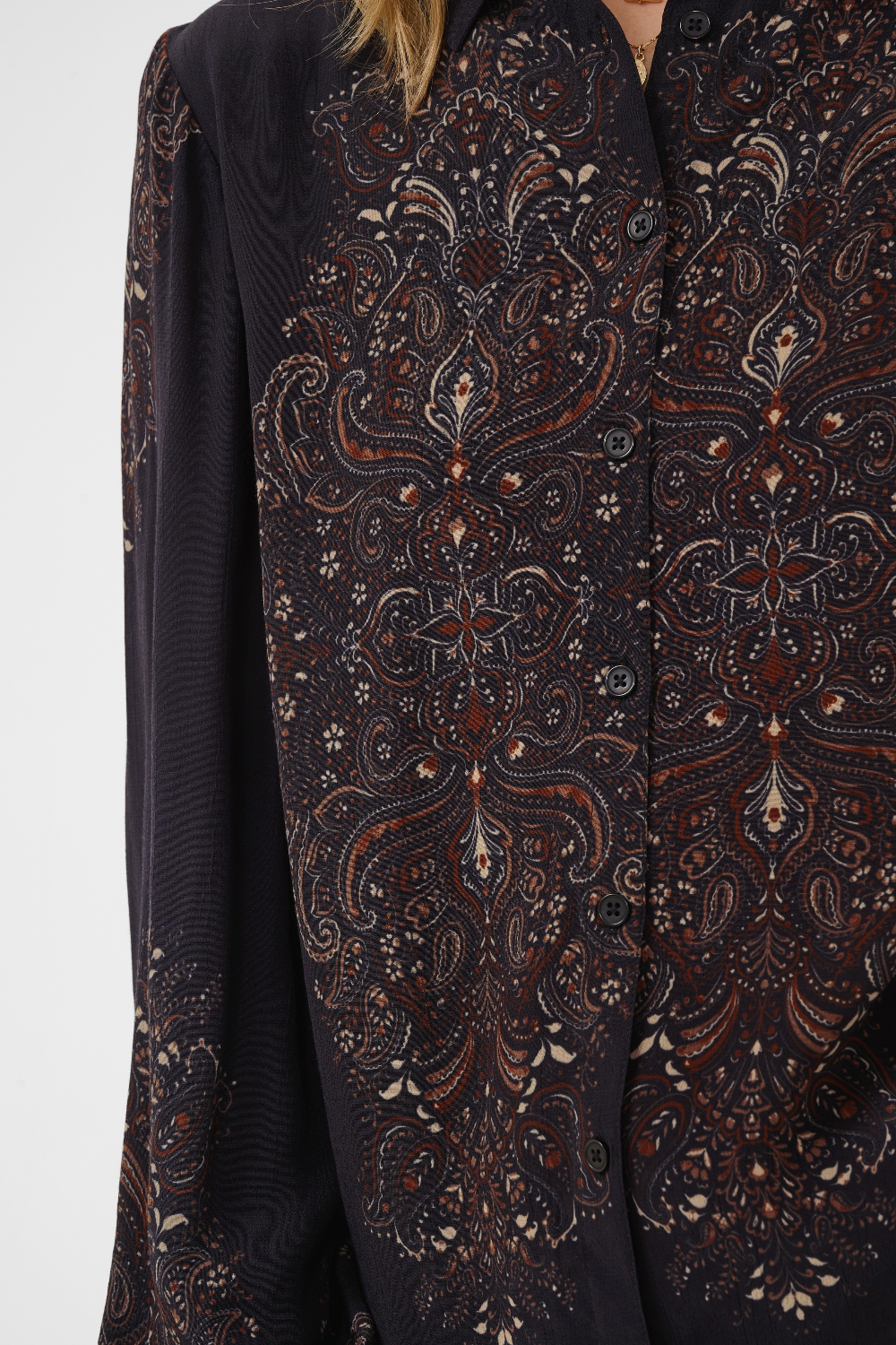 Close-up of a long-sleeve button-front blouse with an intricate paisley and floral pattern in dark brown, rust red, and cream on a black base, worn by a model.