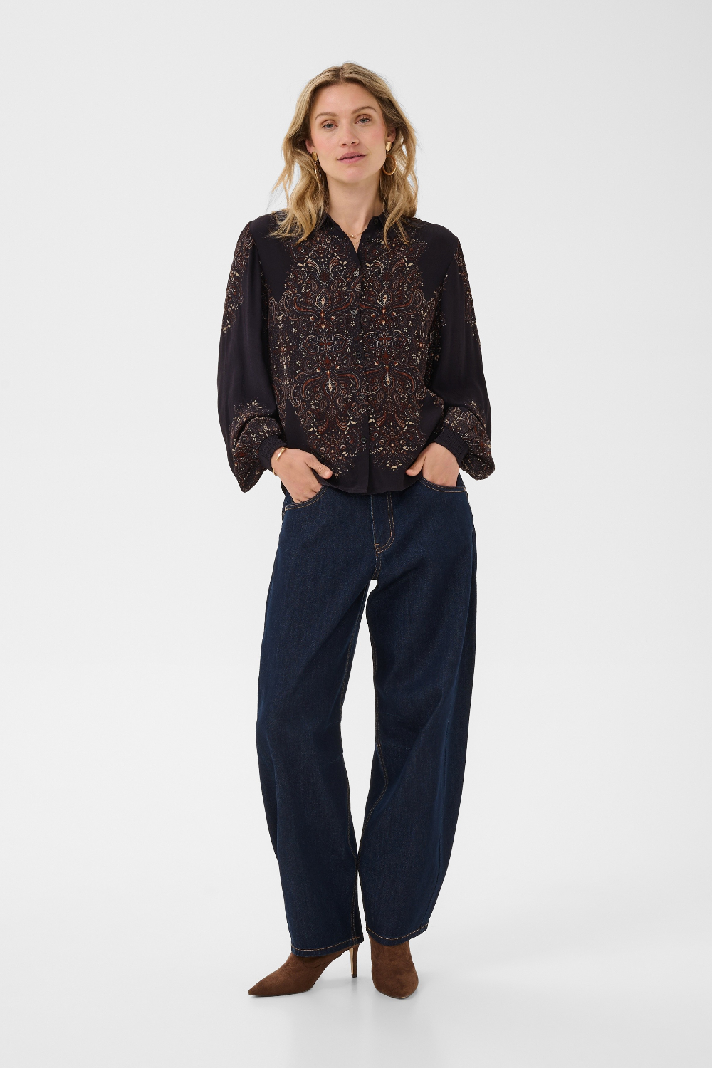 A woman modeling a black long-sleeve button-up blouse with intricate brown and gold floral embroidery across the chest and sleeves, paired with dark blue wide-leg jeans and brown heeled shoes, against a plain white studio background.