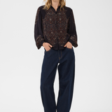 A woman modeling a black long-sleeve button-up blouse with intricate brown and gold floral embroidery across the chest and sleeves, paired with dark blue wide-leg jeans and brown heeled shoes, against a plain white studio background.