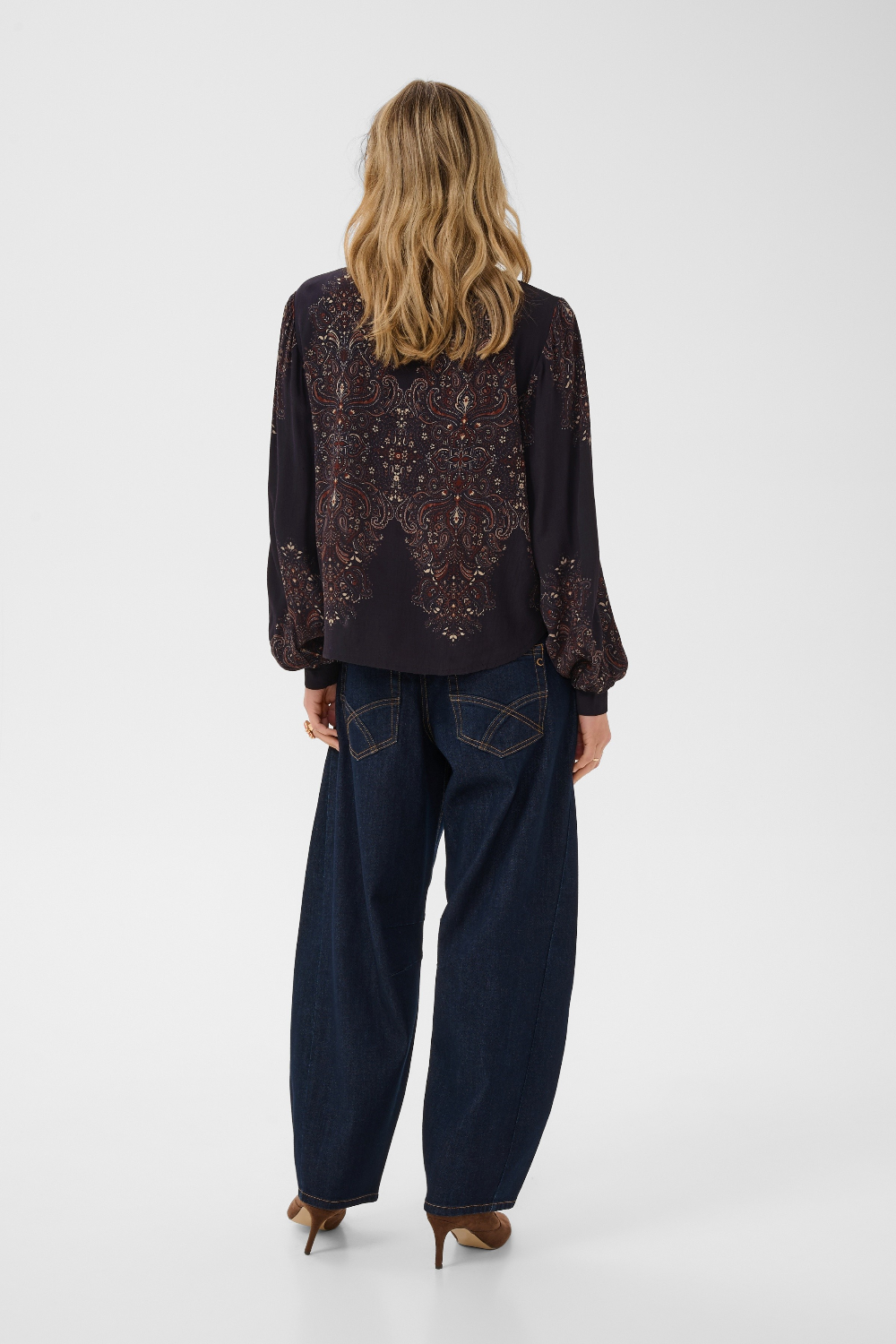 A woman viewed from the back wearing a dark long-sleeve blouse with ornate embroidered detailing across the back and sleeves, paired with dark wide-leg jeans and brown heels, against a plain light gray studio background.