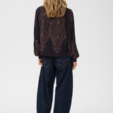 A woman viewed from the back wearing a dark long-sleeve blouse with ornate embroidered detailing across the back and sleeves, paired with dark wide-leg jeans and brown heels, against a plain light gray studio background.