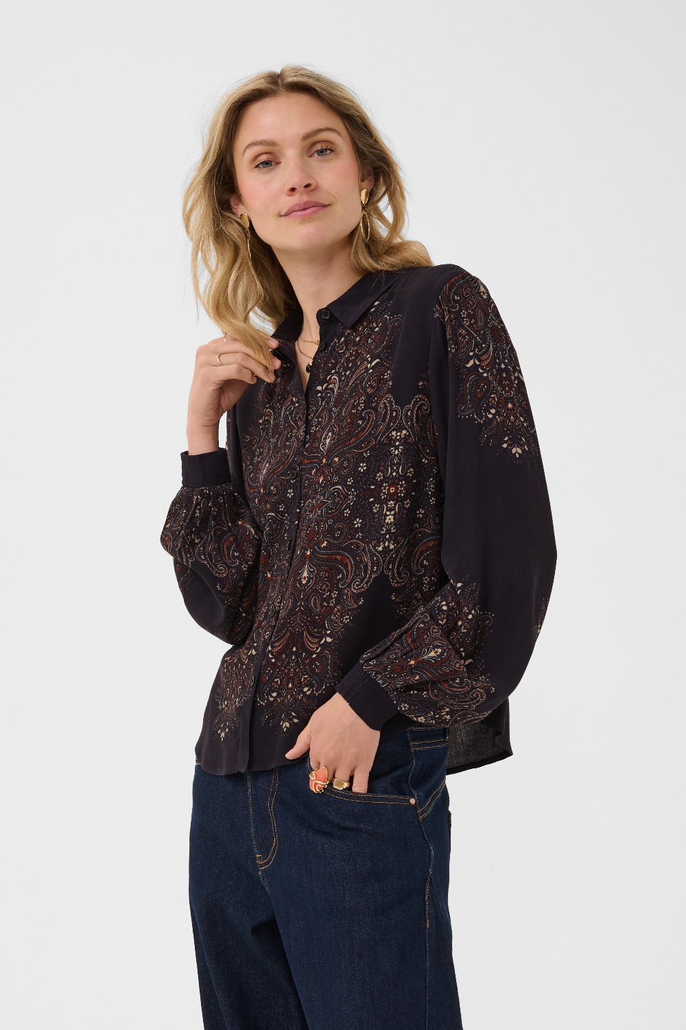 A woman modeling a dark long-sleeve button-up blouse with intricate paisley embroidery, paired with dark blue jeans against a plain light gray studio background.