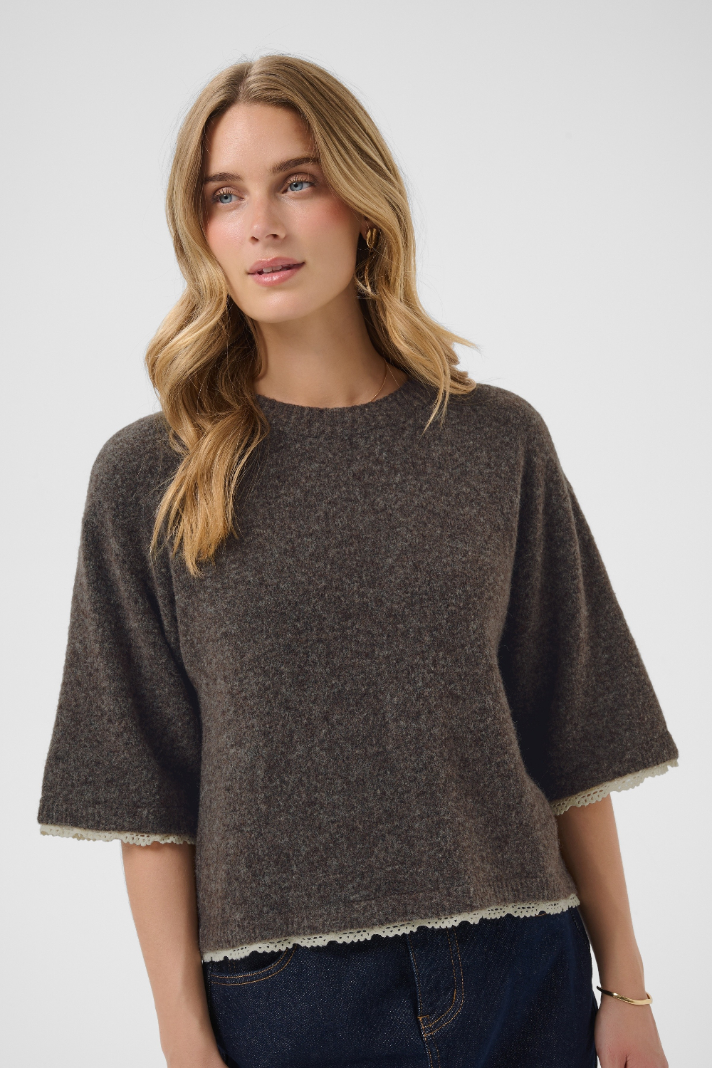 A woman wearing a dark brown textured knit sweater with wide three-quarter sleeves and a scalloped cream trim, paired with dark blue jeans, against a light gray studio background.