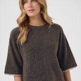 A woman wearing a dark brown textured knit sweater with wide three-quarter sleeves and a scalloped cream trim, paired with dark blue jeans, against a light gray studio background.