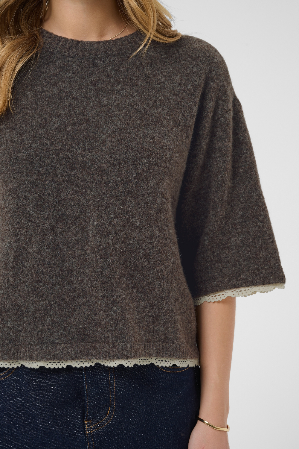 A close-up of a person wearing a dark brown knit sweater with textured heathered detailing and a scalloped cream lace trim at the hem and sleeve cuffs, paired with dark blue jeans.
