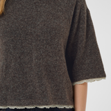 A close-up of a person wearing a dark brown knit sweater with textured heathered detailing and a scalloped cream lace trim at the hem and sleeve cuffs, paired with dark blue jeans.