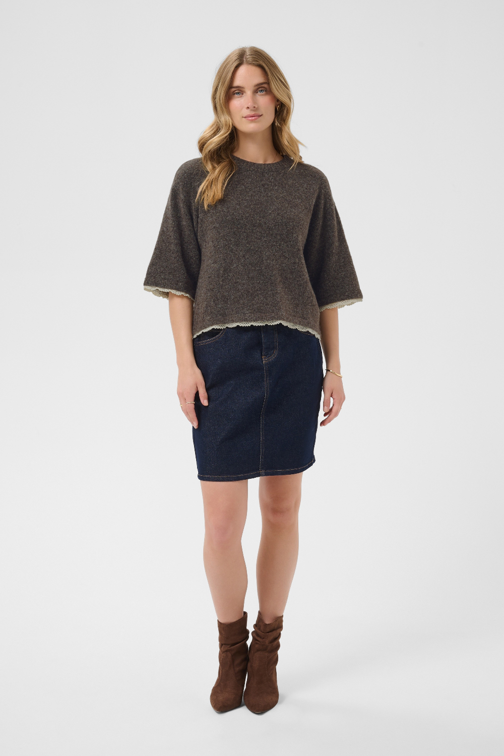 A woman modeling a dark brown short-sleeved knit sweater with a light hem, paired with a dark blue denim mini skirt and brown suede ankle boots, against a light gray studio background.