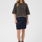 A woman modeling a dark brown short-sleeved knit sweater with a light hem, paired with a dark blue denim mini skirt and brown suede ankle boots, against a light gray studio background.