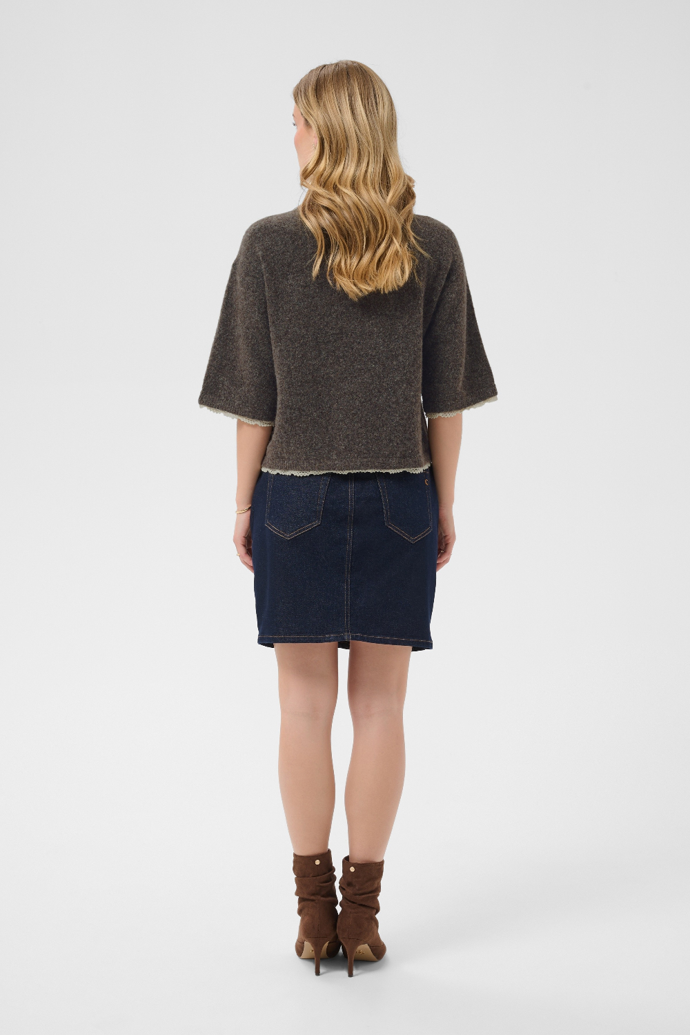 A woman is shown from the back wearing a dark brown textured short-sleeve sweater and a dark blue denim mini skirt, paired with brown ankle boots, against a light gray studio background. 