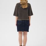 A woman is shown from the back wearing a dark brown textured short-sleeve sweater and a dark blue denim mini skirt, paired with brown ankle boots, against a light gray studio background. 