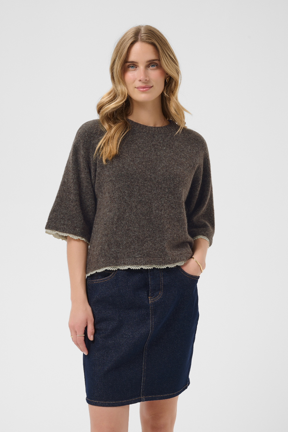 A woman modeling a dark brown heathered knit sweater with a light coloured hem and elbow-length sleeves, paired with a dark blue denim skirt, against a light gray studio background.